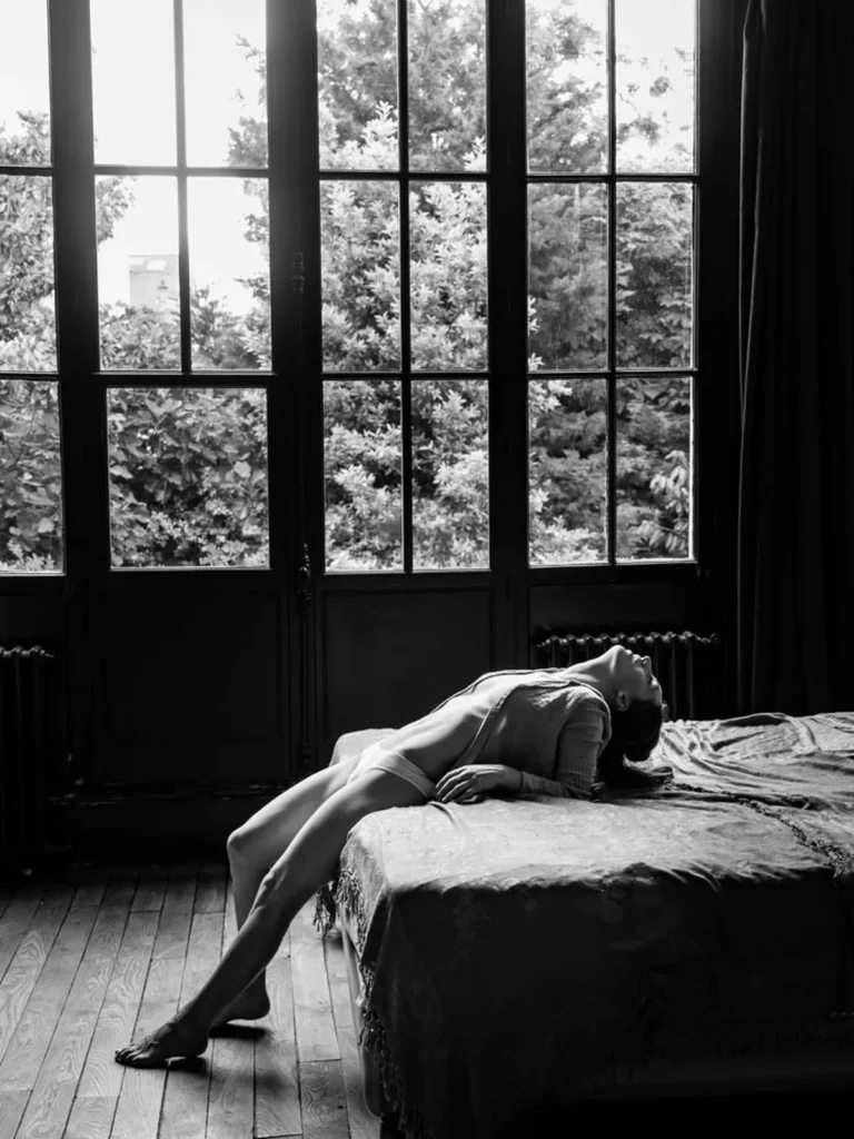 Portrait en noir et blanc d'une femme allongée sur un lit devant une grande fenêtre photographiée par Antoine Cassane.